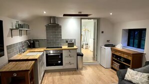 Una cocina moderna con una isla central, armarios de madera y horno empotrado. Hay una zona de comedor con mesa de madera y sofá gris.