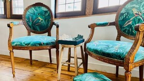 Une pièce comprenant deux fauteuils en bois richement décorés, recouverts d’un tissu turquoise, une petite table en bois sur laquelle est posé un jeu d’échecs, et une fenêtre donnant sur l’extérieur.
