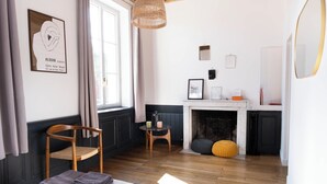 Een kamer met een open haard, een houten stoel, een kleine tafel met een lamp en een lijst met een kunstwerk aan de muur.