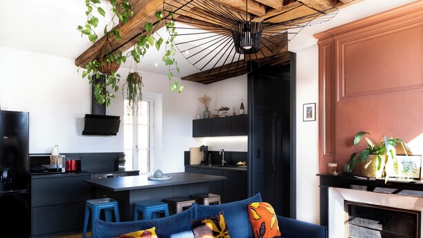 Een moderne keuken met een centraal eiland, een blauwe bank voorzien van kleurrijke kussens, en een hangende plant.