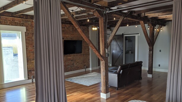 A spacious loft with exposed wooden beams, a large window with curtains, a flat-screen TV mounted on a brick wall, and a cozy seating area with a sofa and a coffee table.