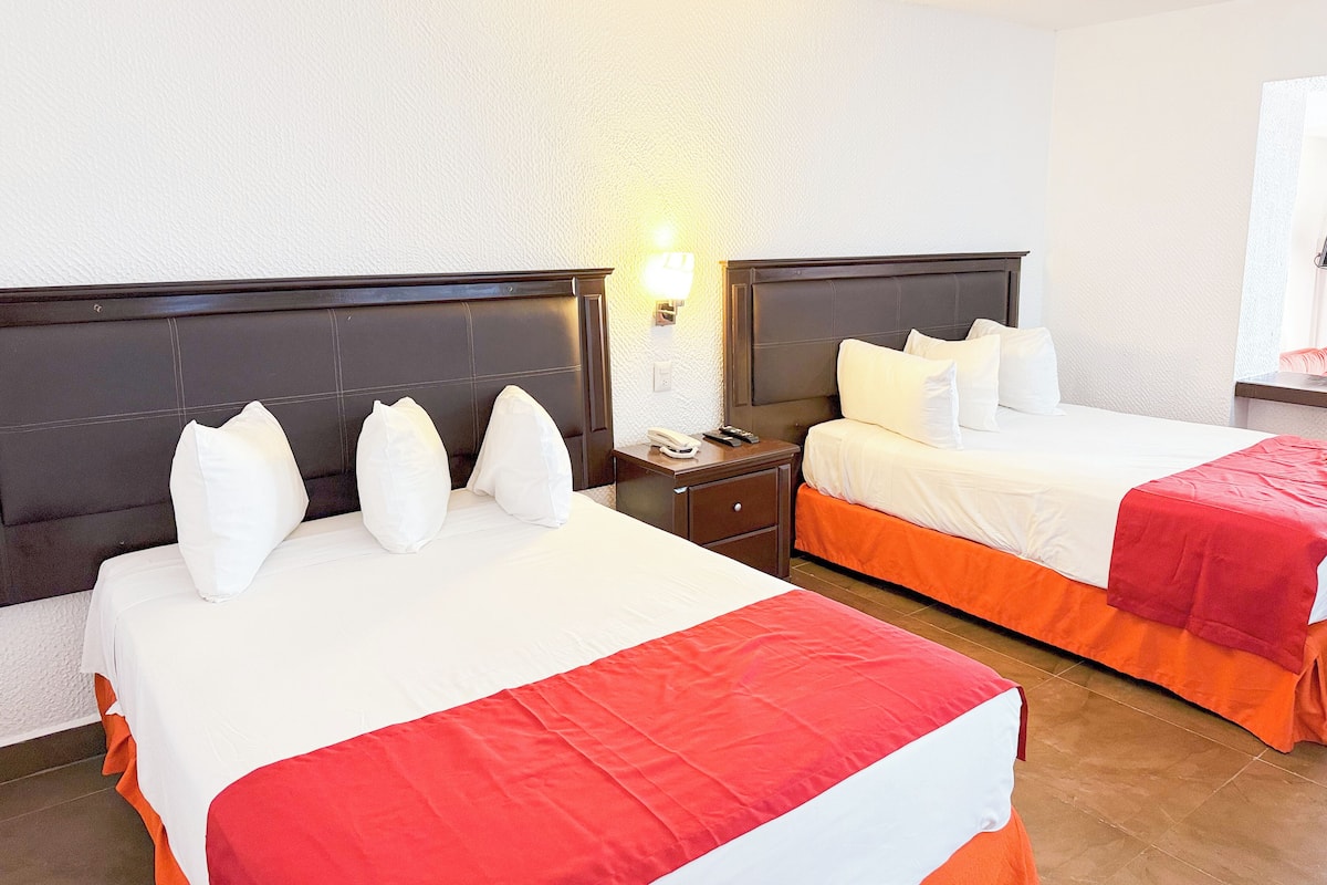 A hotel room with two beds, each with white bedding and red accents, a nightstand with a phone, and a wall-mounted light fixture.