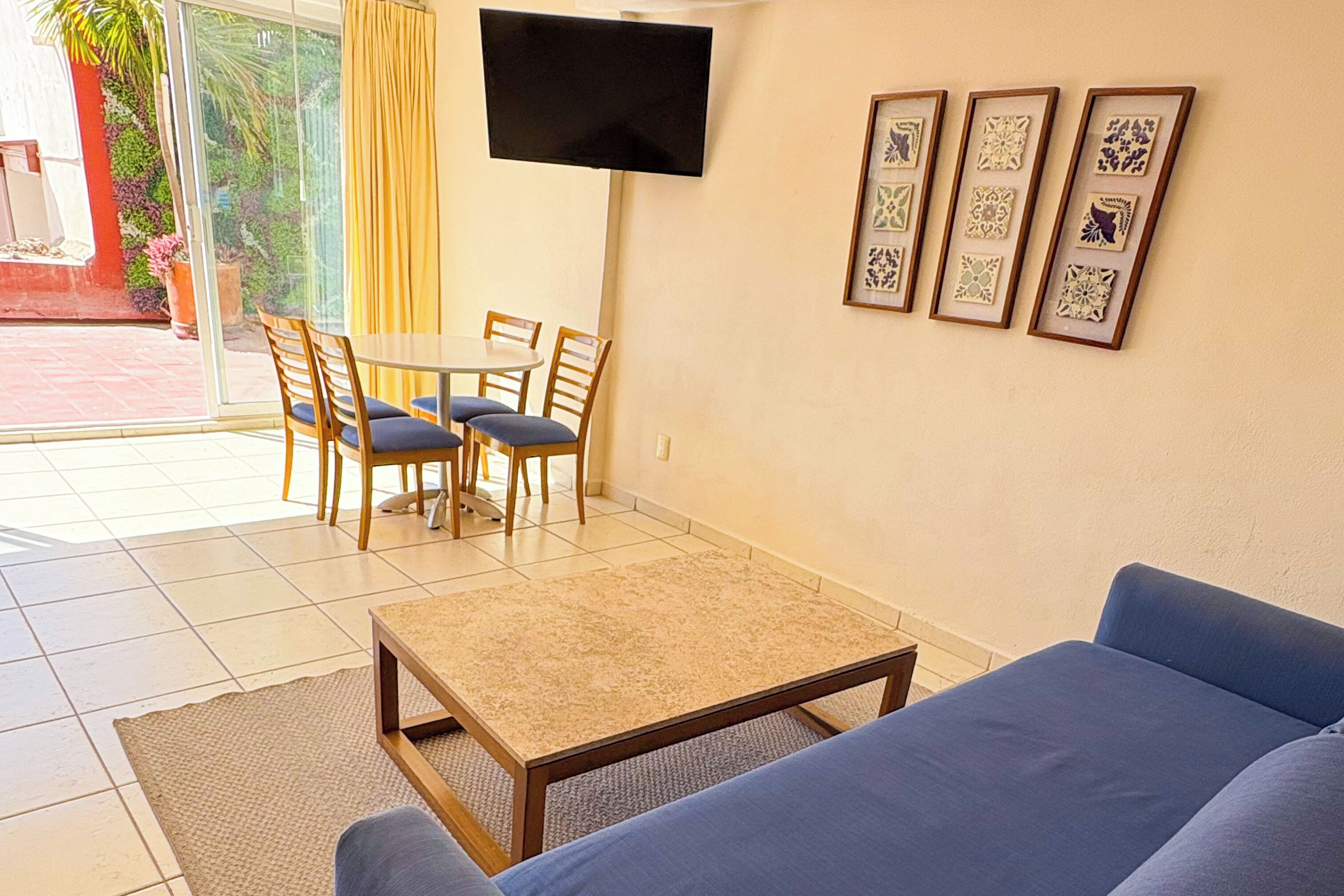 A living room with a sofa, a coffee table, a TV mounted on the wall, and a dining area with chairs and a table.
