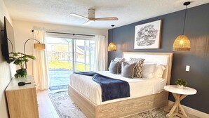 Une chambre avec un grand lit, un ventilateur de plafond, une table de chevet, une petite table ronde et une vue sur l’extérieur, visible à travers une porte vitrée.
