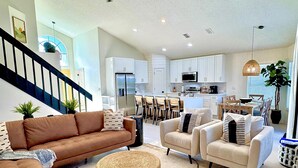 Un vaste espace de vie comprenant un canapé marron, des fauteuils beige et un pouf en osier. On aperçoit à l’arrière-plan une cuisine avec des meubles blancs et un coin repas avec des chaises en bois.