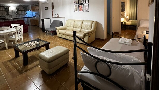 Uma sala de estar com sofá, banquinho e uma cama com estrutura de metal. Há uma mesa de jantar com cadeiras e um lustre.