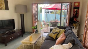 A living room with a TV, sofa, and coffee table. There is a sliding door leading to an outdoor patio with a table and chairs. - North shore room with pool in tropical garden next to beach & Browns Bay village (Auckland)