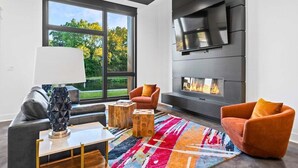 Een moderne woonkamer met een open haard, een bank, twee fauteuils, een salontafel en een groot raam met uitzicht op bomen.
