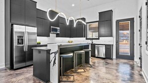 A modern kitchen with a central island, dark cabinetry, stainless steel appliances, and a unique light fixture.