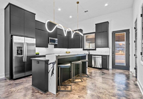 Une cuisine moderne dotée d’un îlot central, de meubles de rangement aux couleurs sombres, d’appareils électroménagers en acier inoxydable et d’un luminaire original.