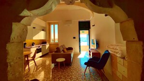 Una sala de estar acogedora con un sofá, una mesita y un sillón. Hay una zona de cocina con mostrador y ventana. El piso tiene un diseño y las paredes son de piedra.