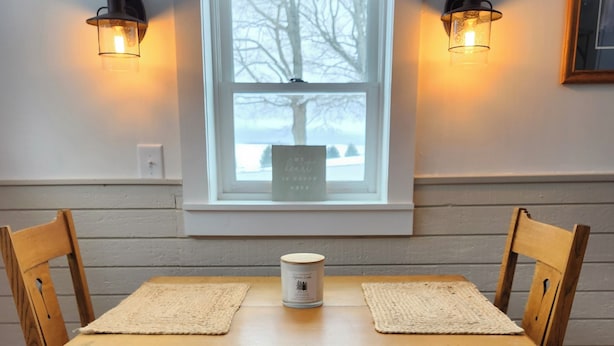 A wooden dining table with two chairs, two wall-mounted light fixtures, a window with a view of trees, and a framed picture on the wall.