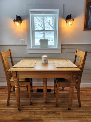 Una mesa de madera con dos sillas, dos lámparas fijadas a la pared, una ventana con vista a árboles y un cuadro enmarcado en la pared.