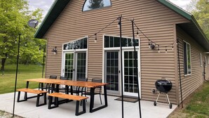A two-story house with a sloped roof, a covered patio with a wooden table and benches, and a barbecue grill outside.