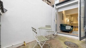 Un pequeño espacio exterior con mesa y dos sillas, una puerta corrediza de vidrio que da a una sala de estar y una pared blanca.
