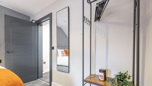 Una habitación de hotel moderna con una puerta gris, un espejo grande, un estante de madera con un tarro y una planta, y una cama con ropa de cama naranja.