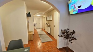 A hallway with a TV mounted on the wall, a dining area with chairs, a plant, and a kitchen area in the background.