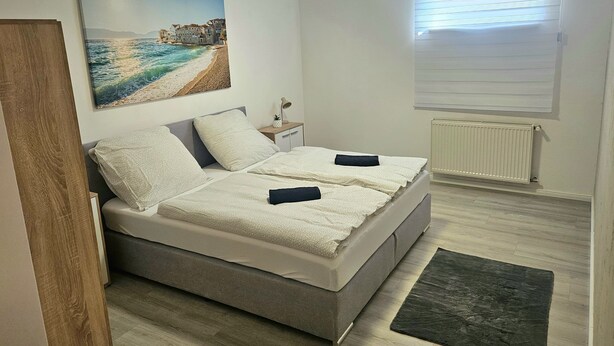 A bedroom with a bed, a nightstand, a window with blinds, and a painting on the wall.