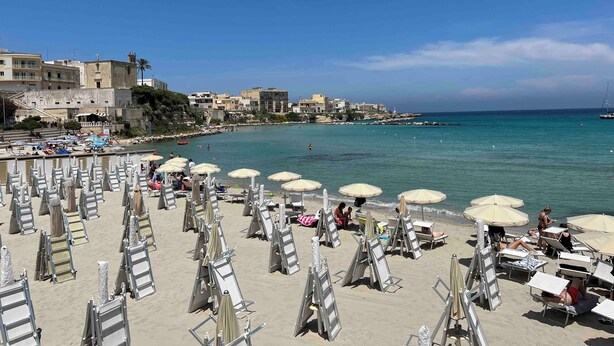 Ein Strand mit Reihen von Liegestühlen und Sonnenschirmen, einem klaren blauen Meer und Gebäuden im Hintergrund.