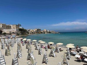A beach with rows of sun loungers and umbrellas, a clear blue sea, and buildings in the background.