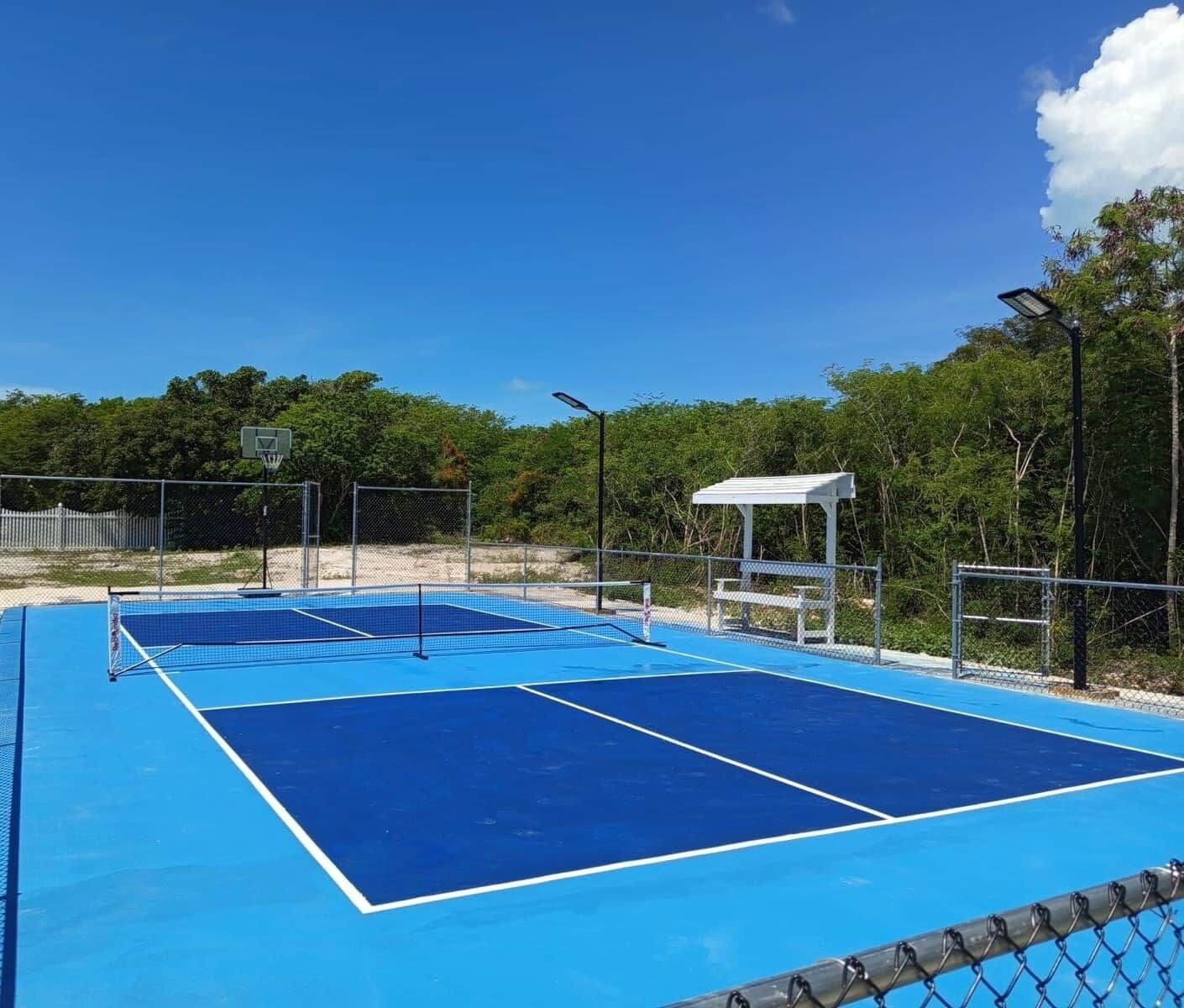 An outdoor tennis court with a blue surface, surrounded by a chain-link fence, and a white pavilion in the background.