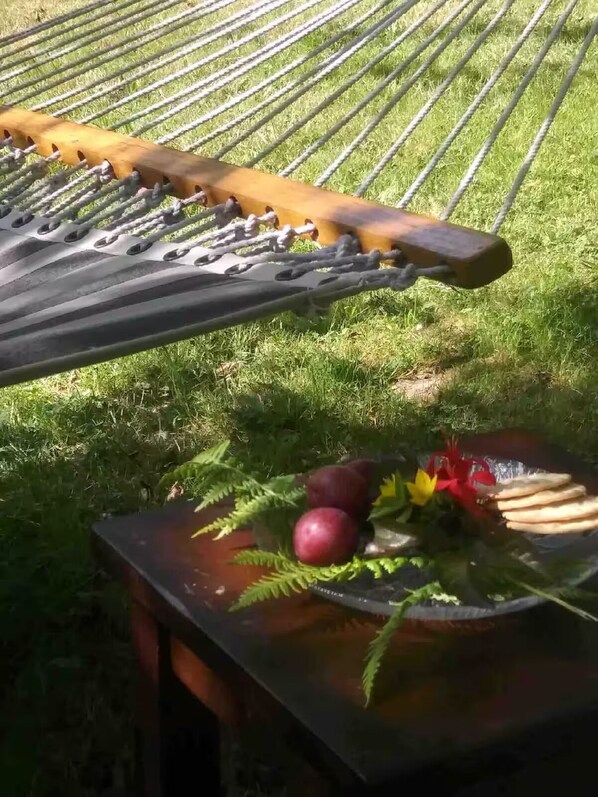 En hängmatta som hänger över en gräsyta, och under den står ett bord med frukt och en blomsteruppsättning.