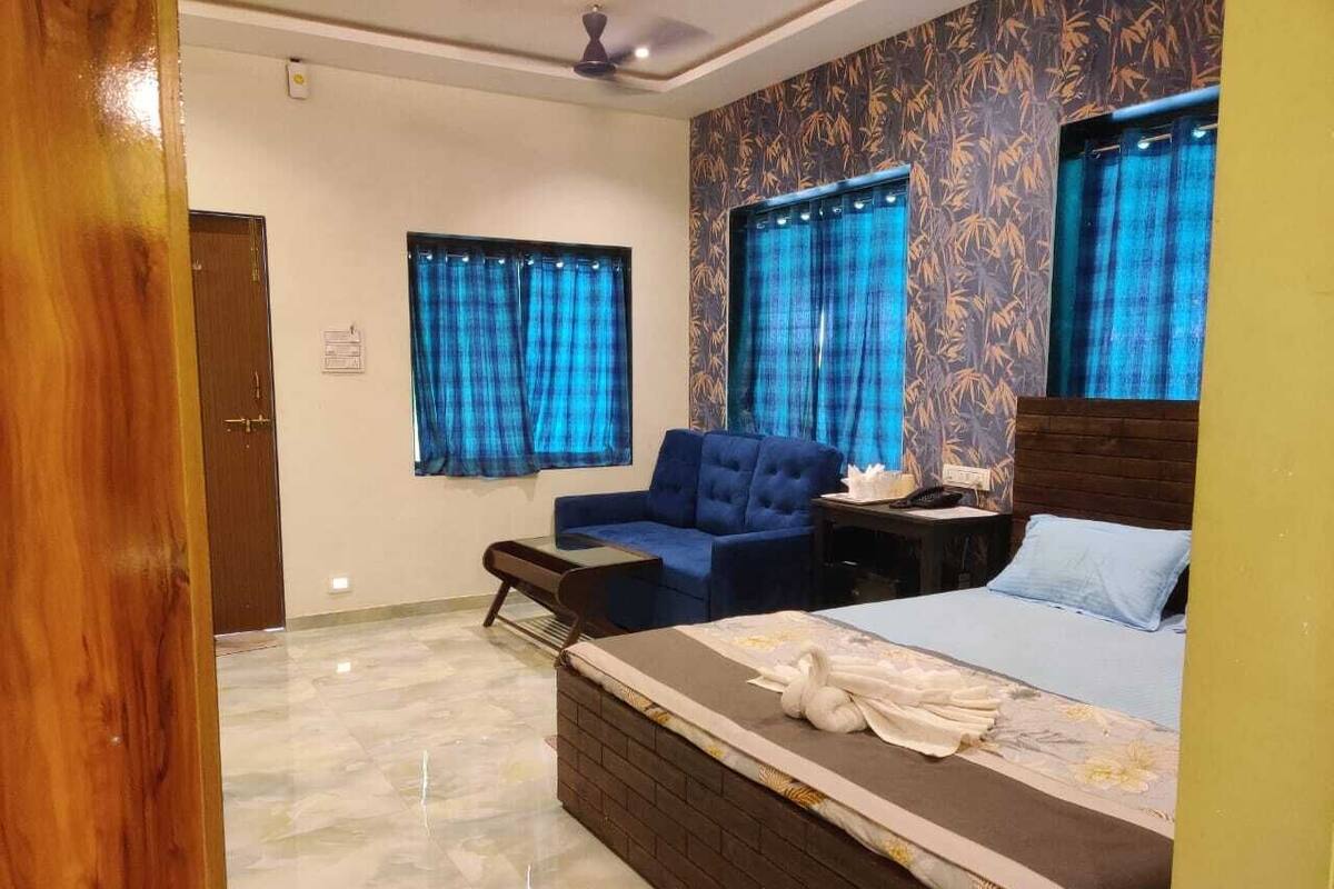Quarto de hotel com uma cama, um sofá azul, uma escrivaninha com telefone e duas janelas com cortinas azuis.