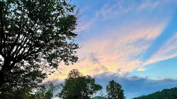 Una casa con terraza, rodeada por un campo cubierto de hierba y árboles, bajo un cielo parcialmente nublado.