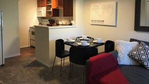 Una cucina compatta con mobili in legno, un forno a microonde e un lavandino. C'è una zona pranzo con un tavolo apparecchiato per quattro, sedie nere e una coperta rossa sul divano.