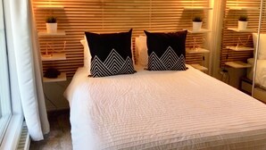 Un letto rifatto con un comofortino bianco e due cuscini neri a motivi geometrici. Si notano due comodini con lampade, una mensola a parete con una pianta e un cartello con la scritta 'Welcome' sopra il letto.