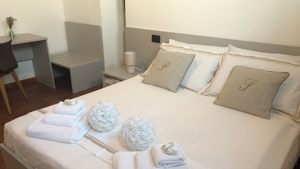A neatly made bed with towels, pillows with initials, a small table with a vase, and a chair. - Il piccolo scrigno  (Lonigo)