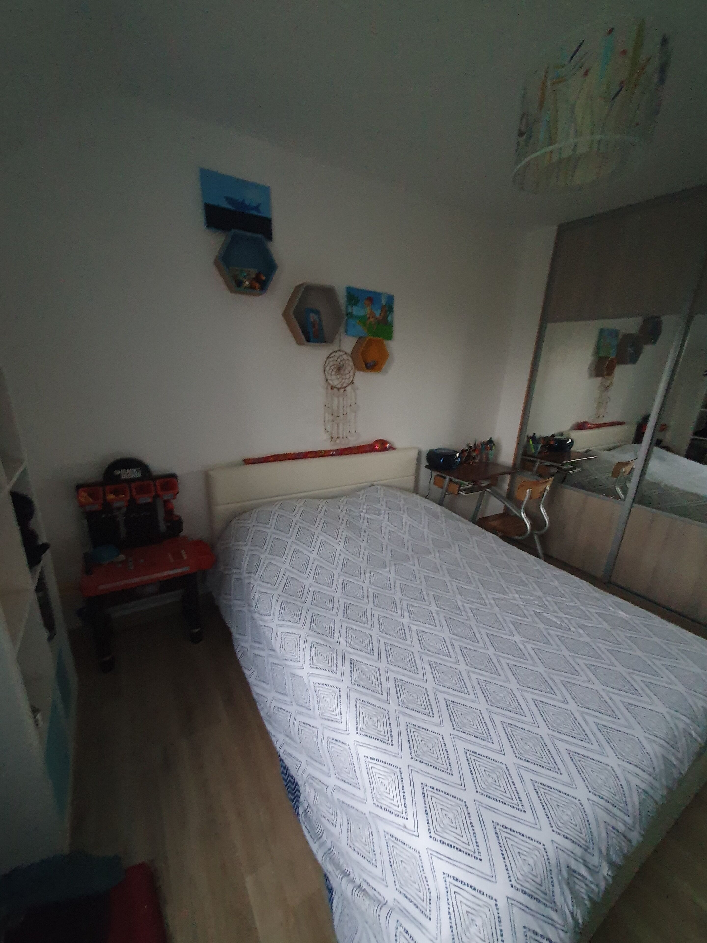 Une chambre à coucher avec une couvre-lit à motifs, un bureau avec une chaise, une commode et des décorations murales, notamment un attrape-rêves et des tableaux encadrés.