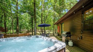 A hot tub in a wooded area with a wooden cabin, outdoor grill, and patio furniture.