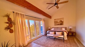 Quarto com cama, mesas de cabeceira, ventilador de teto e uma janela grande com cortinas.