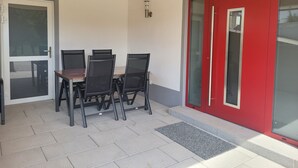 Ein überdachter Patio-Bereich mit Tisch und Stühlen, eine rote Tür und eine Glastür, die nach draußen führt.