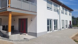 Ein modernes, zweistöckiges Gebäude mit Balkon, roter Tür und überdachter Außenbestuhlung.