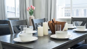 Une table à manger est dressée avec de l’assiette blanche, des bols et une bougie. Des chaises en bois sont disposées autour de la table, et un vase rempli de fleurs se trouve à l’arrière-plan.