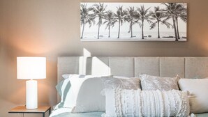 Une chambre à coucher avec un lit, une table de chevet, une lampe et un tableau représentant des palmiers accroché au mur.