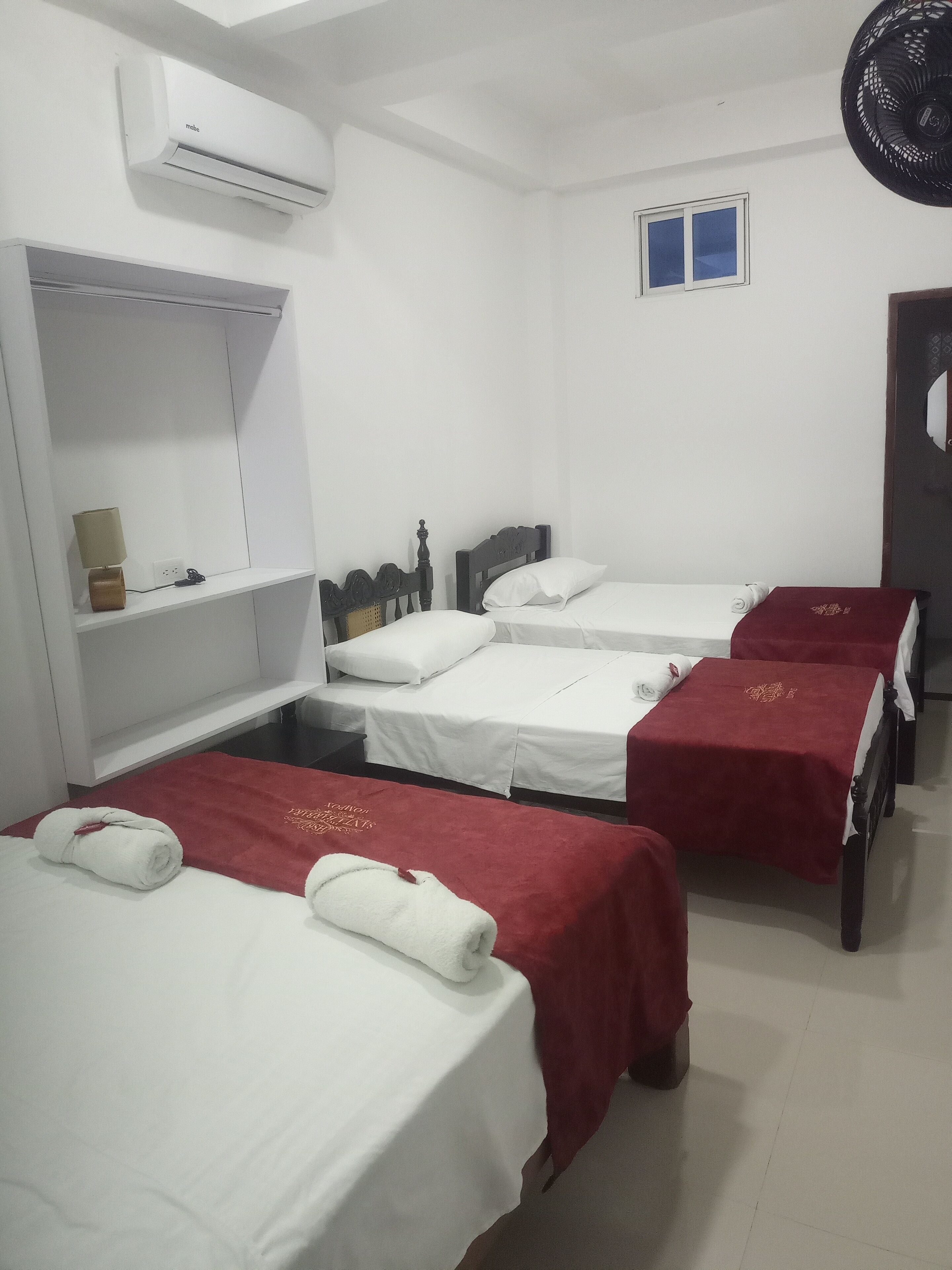 A hotel room with four single beds, each with a red bedspread and white linens. There is a white shelf unit with a lamp and a small decorative item. An air conditioning unit is mounted on the wall above the shelf.