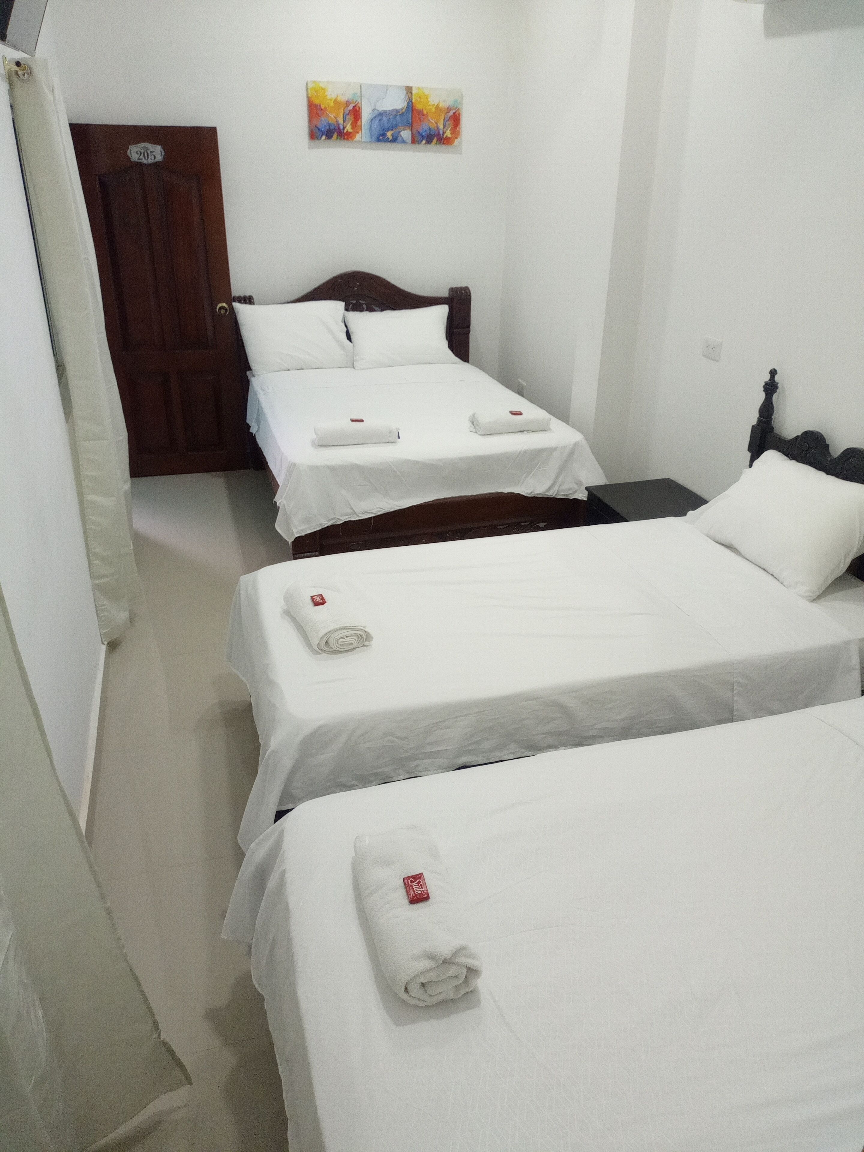 Habitación de hotel con tres camas individuales, cada una con una colcha blanca y una toalla blanca enrollada. Se ve una puerta de madera con una placa de número, una pared con un cuadro abstracto de colores, y una mesita de noche negra con una lámpara.