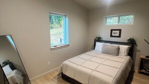 Un dormitorio con una cama, dos ventanas, una mesita de noche y un espejo.