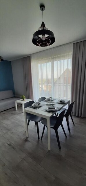 A dining area with a white table set for four, black chairs, a hanging light fixture, and a view of a building through large windows with sheer curtains. - Apartamenty Jasieńczyk (Mielenko)