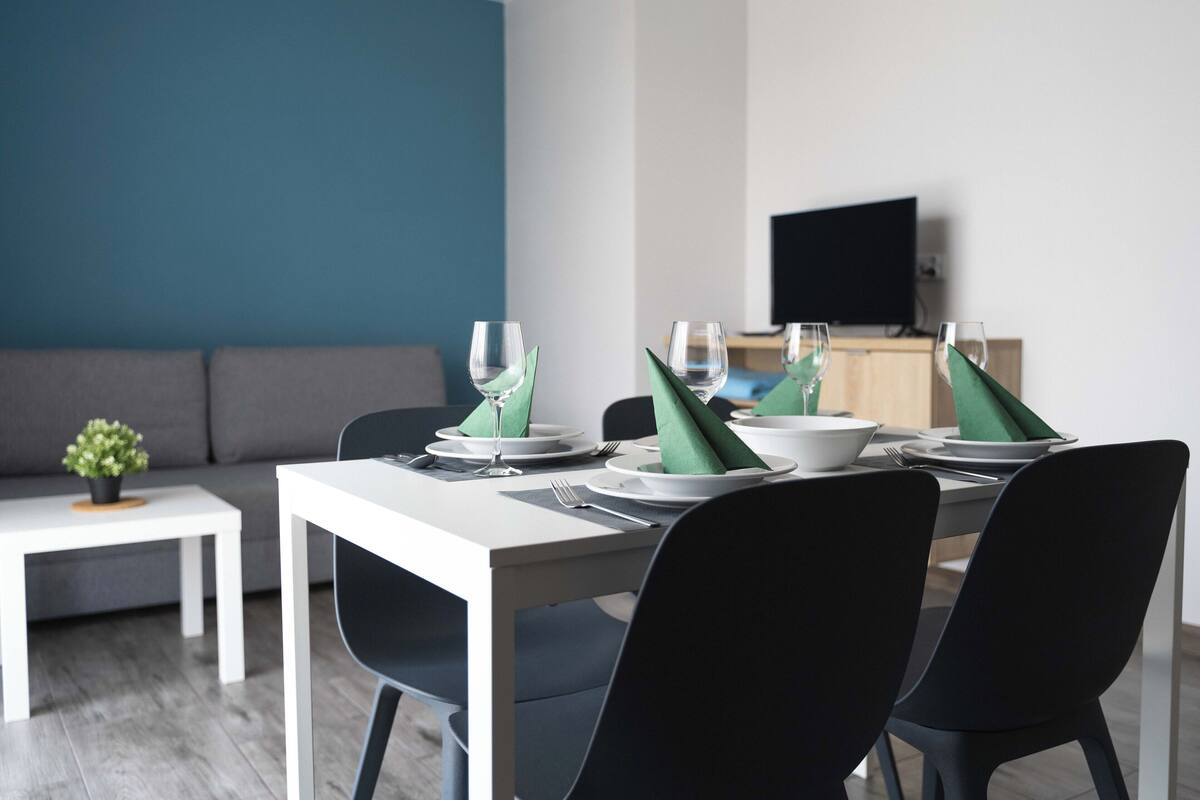 A dining area with a white table set for two, black chairs, and a small side table with a plant.