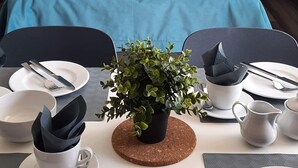 A neatly made bed with blue bedding and white pillows, a small table with a potted plant, and a dining setup with plates, cutlery, and a pitcher. - Apartamenty Jasieńczyk (Mielenko)