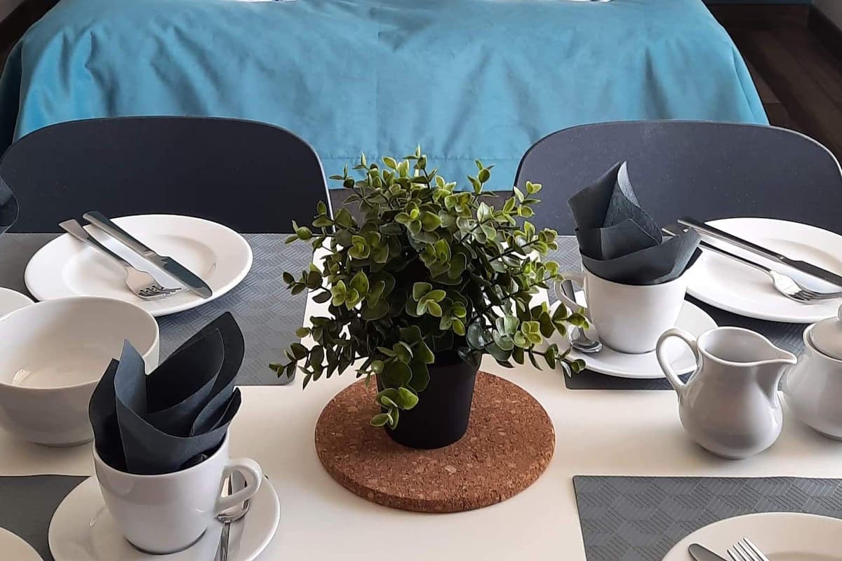 A neatly made bed with blue bedding and white pillows, a small table with a potted plant, and a dining setup with plates, cutlery, and a pitcher.