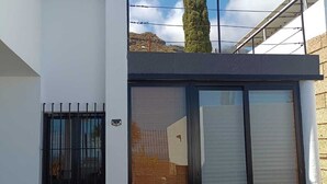 Una moderna casa a due piani con un balcone, un'ampia porta scorrevole in vetro e vista su un albero e delle colline in lontananza.