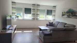 Ein modernes Wohnzimmer mit Sofa, Couchtisch und einem TV-Schrank. Große Fenster mit Jalousien, ein Gemälde an der Wand und Blick auf Grünflächen außerhalb.
