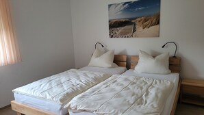Ein Doppelbett mit weißer Bettwäsche und zwei Kissen, ein hölzernes Kopfende, ein Nachttisch mit Lampe und ein Bild einer Strandlandschaft an der Wand.