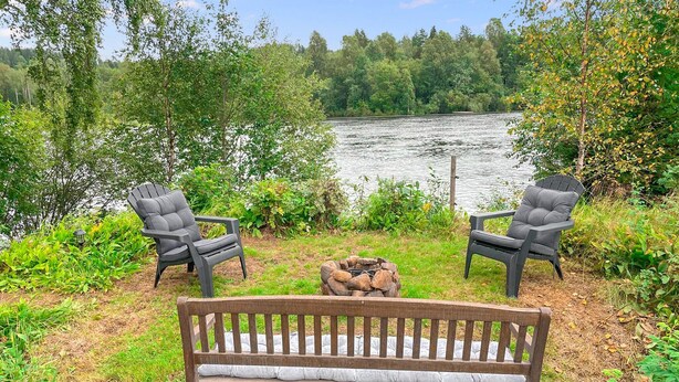 Un banc en bois avec des coussins, deux fauteuils Adirondack et un foyer, le tout installé dans un cadre idyllique au bord du lac.