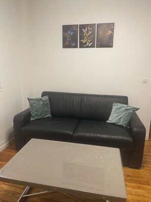 Een woonkamer met een zwarte leren bank, twee gekleurde kussens, een witte salontafel en drie ingelijste kunstwerken aan de muur.